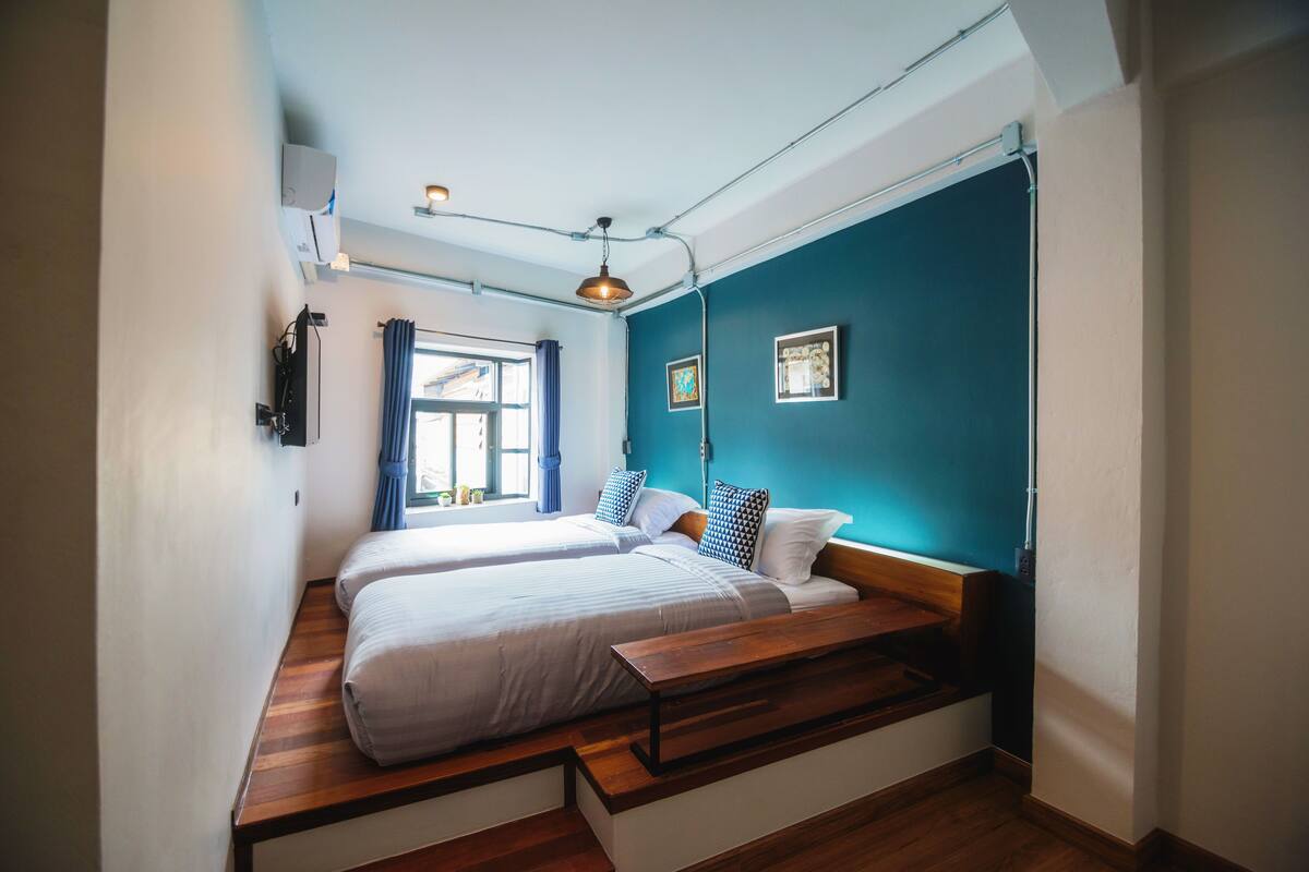 A room with two beds, a wooden bench, a window with curtains, and a ceiling light fixture.