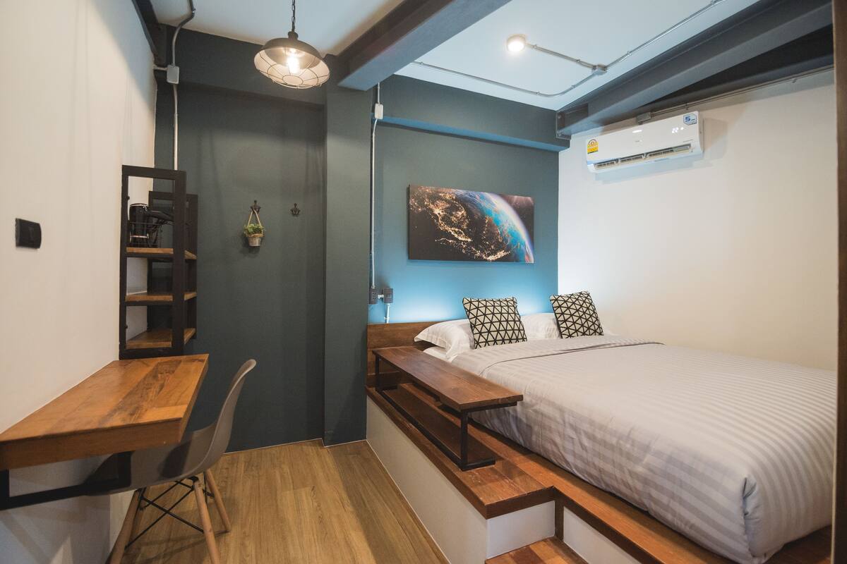 A compact bedroom with a bed, a desk, a chair, a hanging light fixture, and a wall-mounted air conditioner.