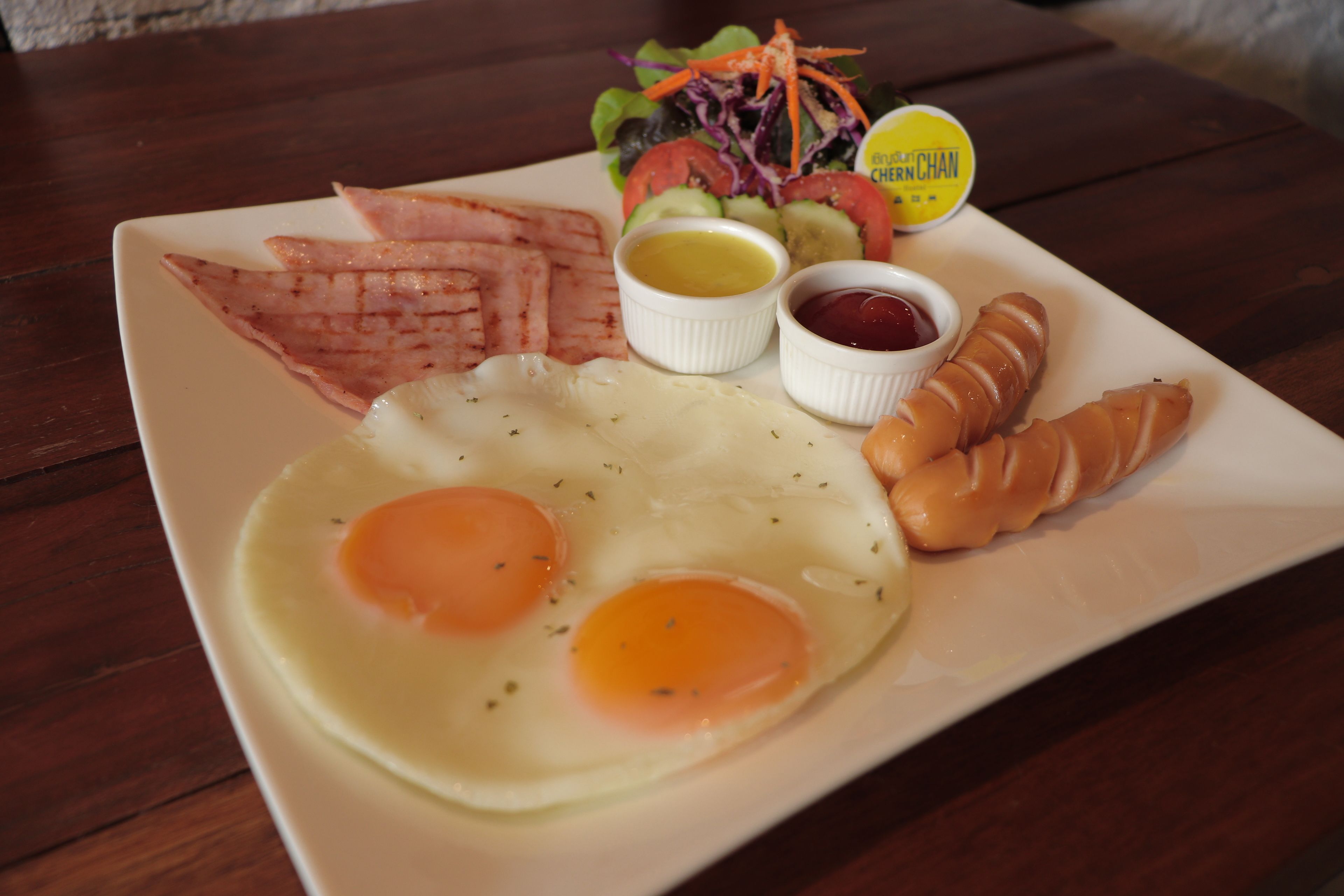 A breakfast plate with eggs, bacon, a croissant, a salad, and two small containers of condiments.