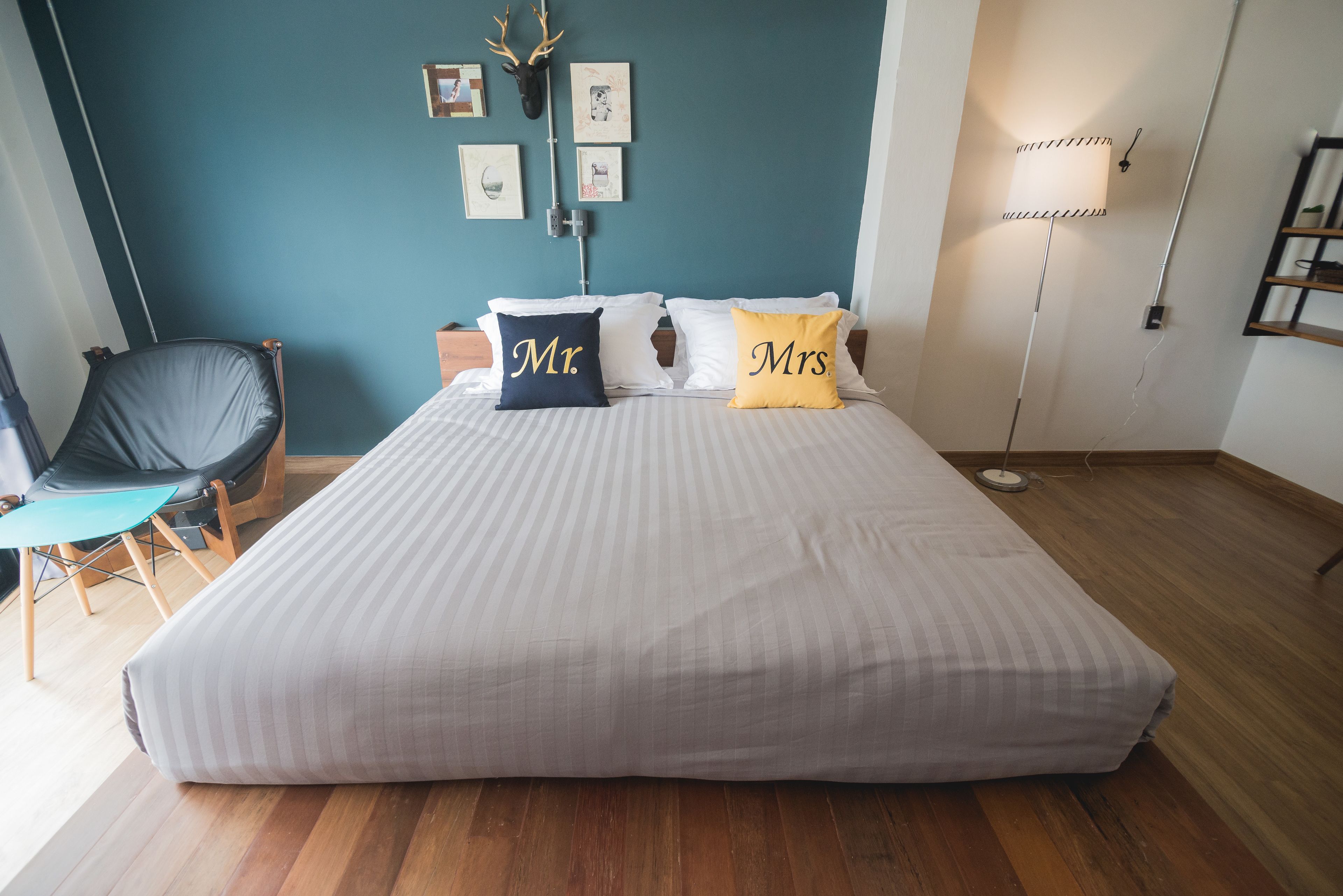 Una cama bien hecha con almohadas que dicen 'Sr.' y 'Sra.' en una habitación con paredes color azul verdoso, piso de madera y una silla con cojín azul verdoso.