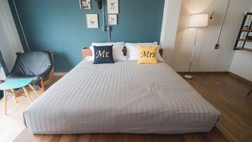 Una cama bien hecha con almohadas que dicen 'Sr.' y 'Sra.' en una habitación con paredes color azul verdoso, piso de madera y una silla con cojín azul verdoso.