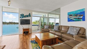 A spacious living room with a leather sofa, a wooden coffee table, and a TV mounted on the wall. A large window offers a view of a body of water and a beach. - BAYFRONT! AUGUST 8 - 10 OPEN AMAZING VIEWS & LOCATION WALK TO FOOD, SHOPS, OCEAN (San Diego)