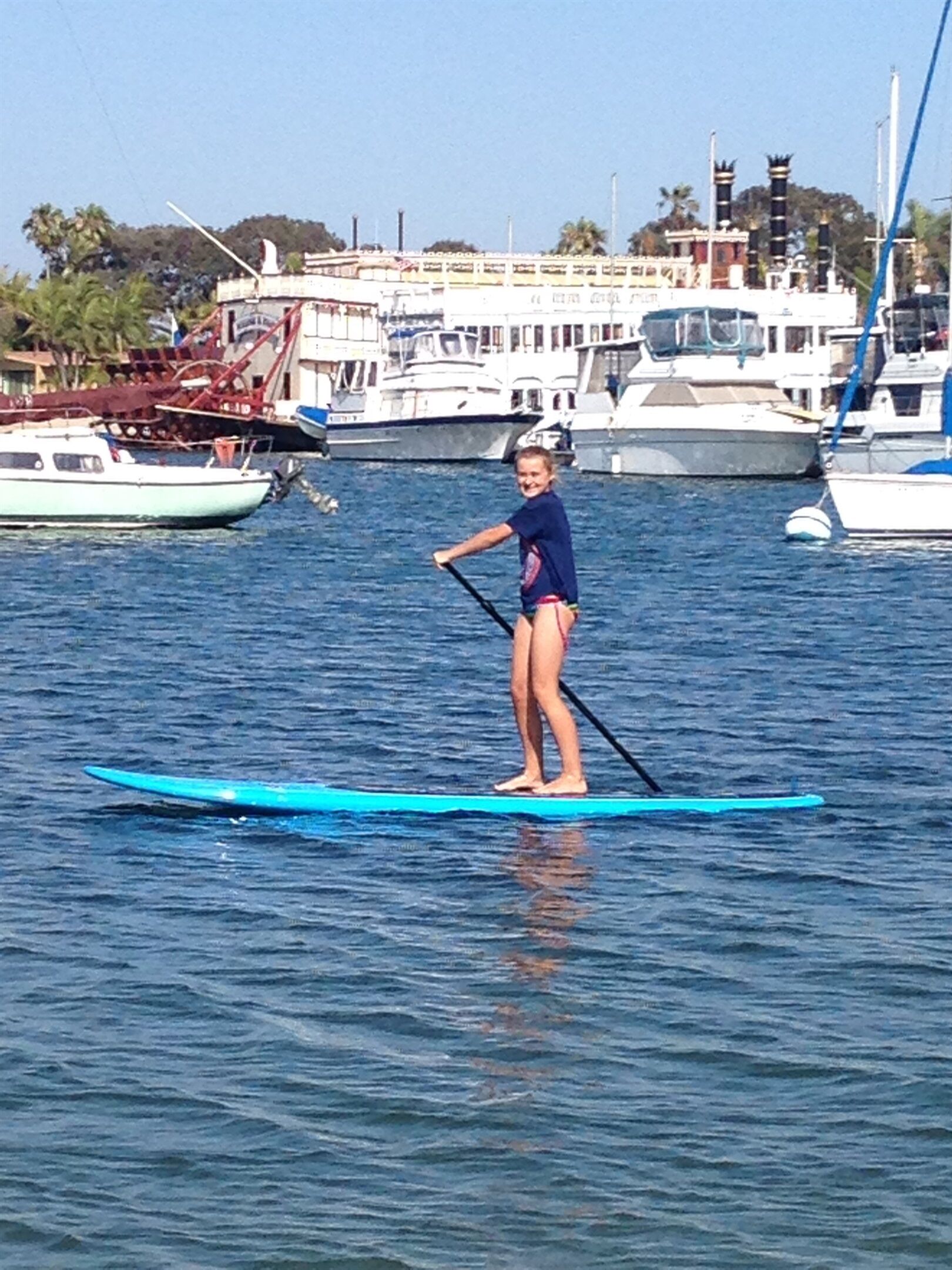 Una persona de pie en una tabla de paddleboard en un puerto deportivo, con barcos y edificios al fondo.