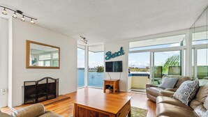 A spacious living room with a large window offering a view of the sea, a wooden coffee table, a leather sofa, and a flat-screen TV mounted on the wall. - BAYFRONT! AUGUST 8 - 10 OPEN AMAZING VIEWS & LOCATION WALK TO FOOD, SHOPS, OCEAN (San Diego)