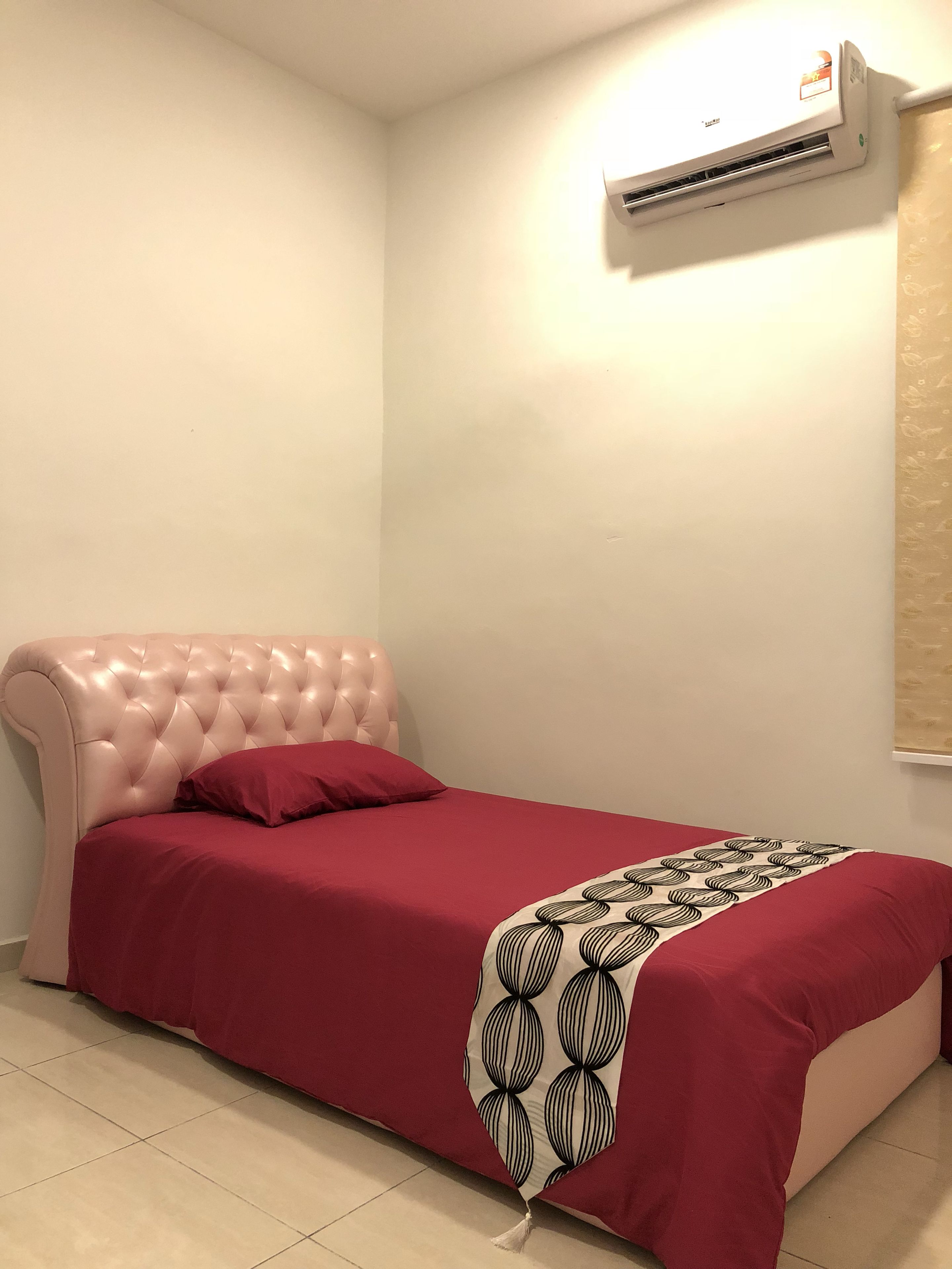 A single bed with a tufted headboard, a red bedspread, and a patterned throw. An air conditioning unit is mounted on the wall above the bed.