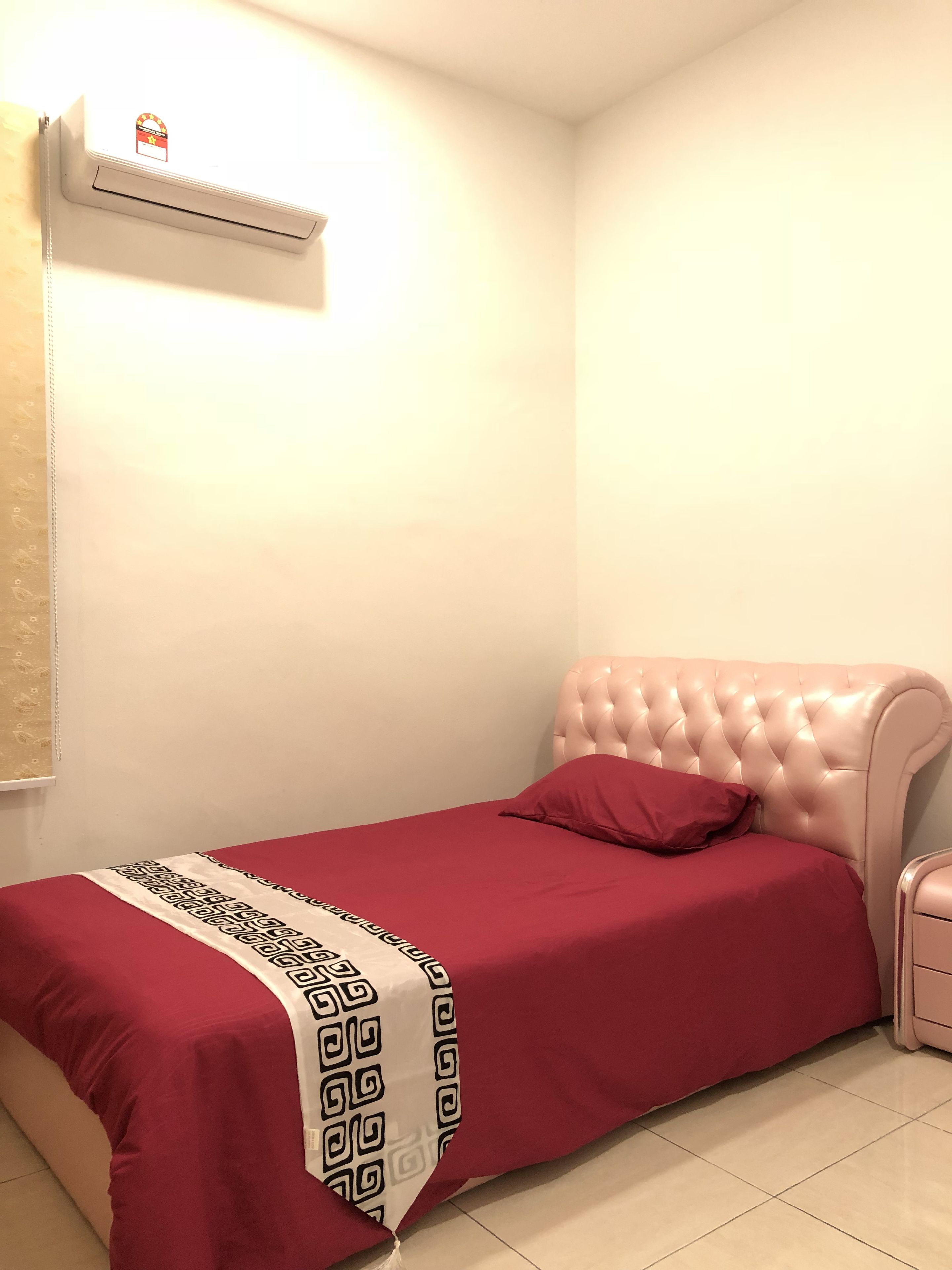 A small, neatly made bed with a red bedspread and a decorative pillow, a white air conditioning unit on the wall, and a white nightstand with a drawer.