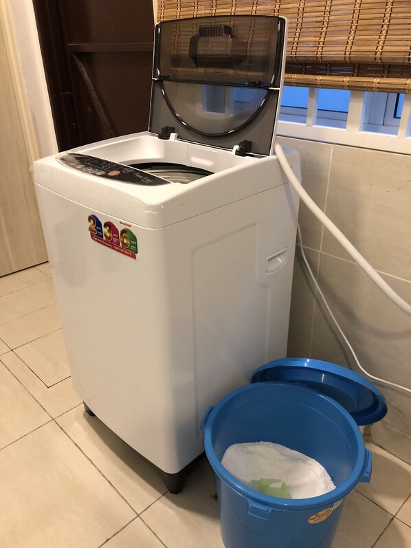 A compact washer and dryer unit with a control panel, placed next to a blue trash bin.