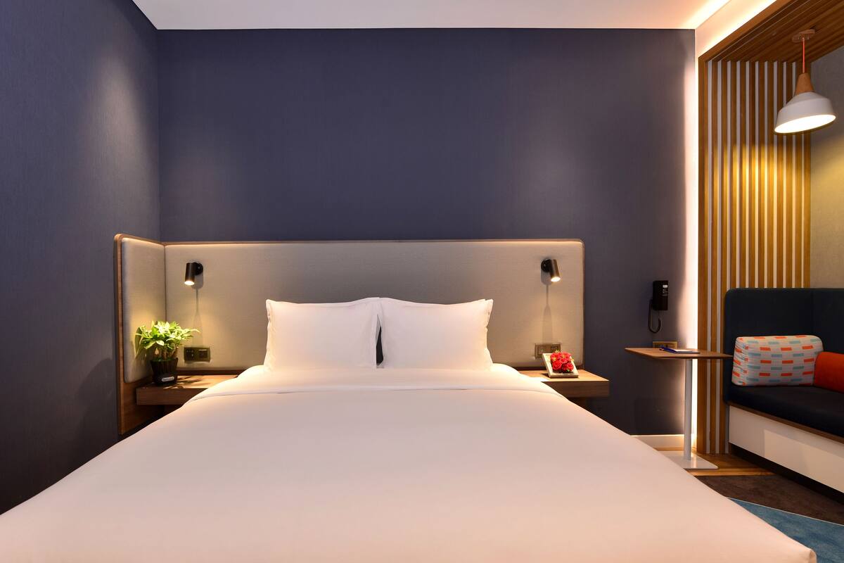 A modern hotel room with a large bed, bedside lamps, a small table with a plant, and a sofa with a patterned cushion.