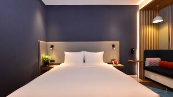 Una habitación de hotel moderna con una cama grande, lámparas de noche, una mesita con una planta y un sofá con un cojín estampado.