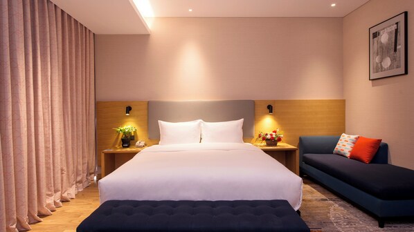 Ein modernes Hotelzimmer mit einem großen Bett, einem Nachttisch mit Lampe, einem kleinen Tisch mit Blumenvase, einer Couch mit Kissen und einem gerahmten Bild an der Wand.