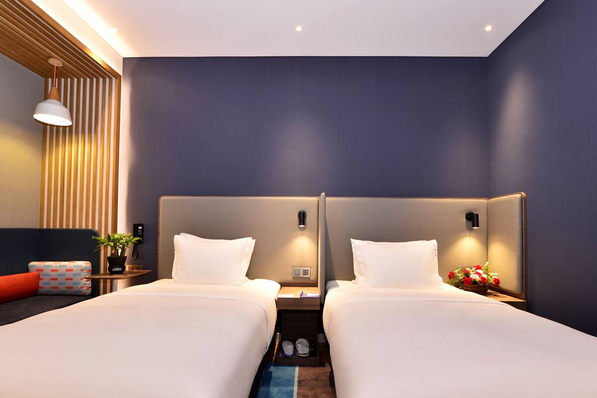 A modern hotel room with two beds, a bedside table with a lamp, a small plant, and a vase of flowers.