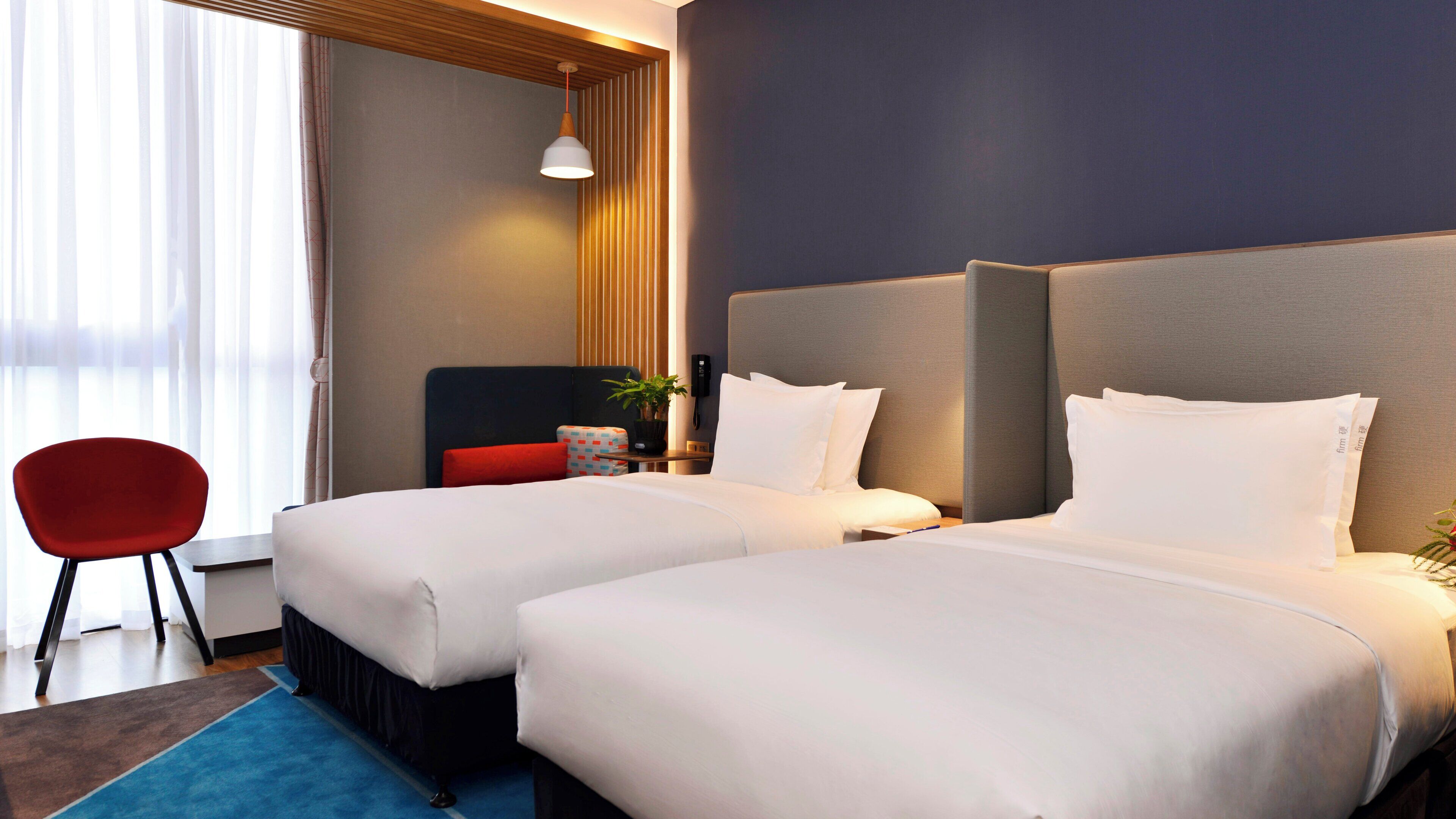 Habitación de hotel con dos camas, una silla roja, una mesita de noche con lámpara y una ventana con cortinas.