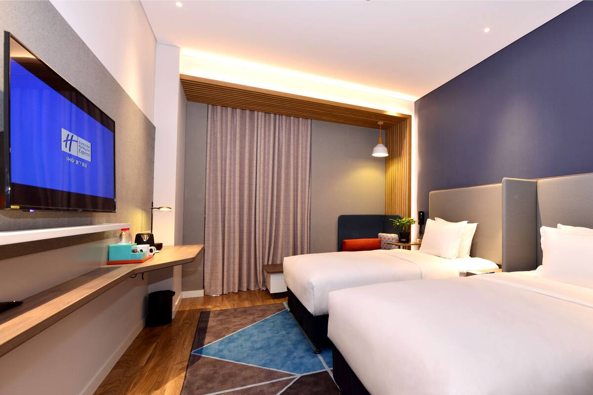 A hotel room with two beds, a TV on a shelf, a desk with a lamp, a small table with a plant, and a window with curtains.