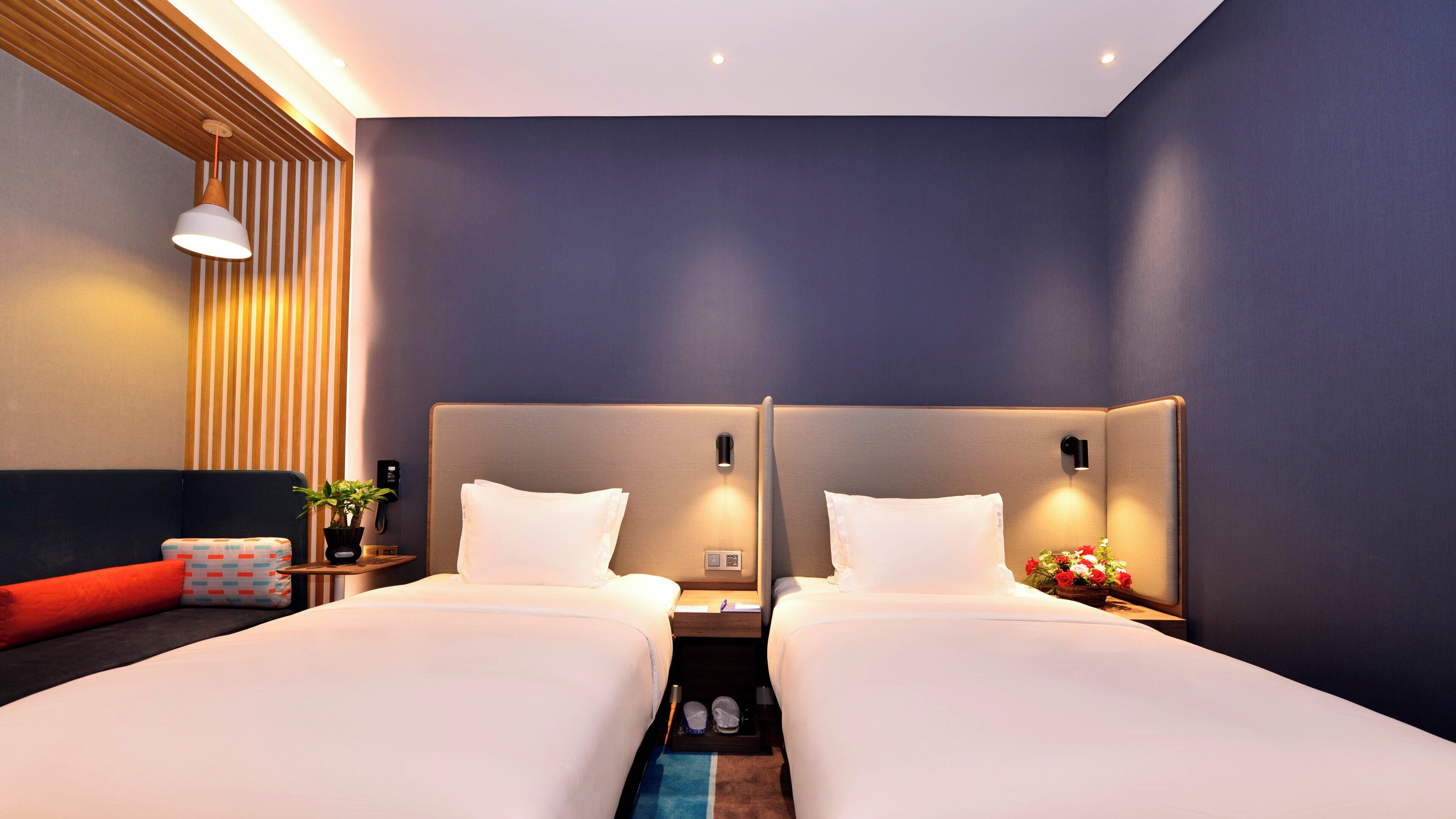 A hotel room with two beds, a bedside table with a lamp, a small table with a plant, and a wall-mounted phone.