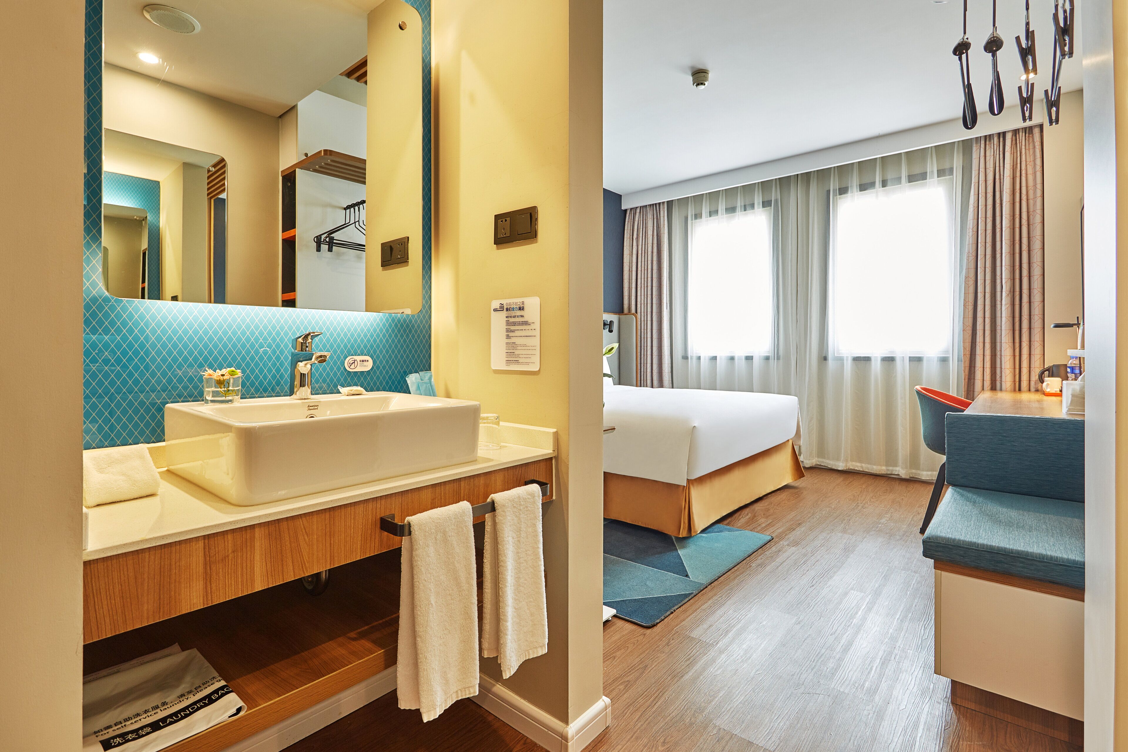 A hotel room with a large bathroom sink, a mirror, a bed, a bedside table, and a window with curtains.