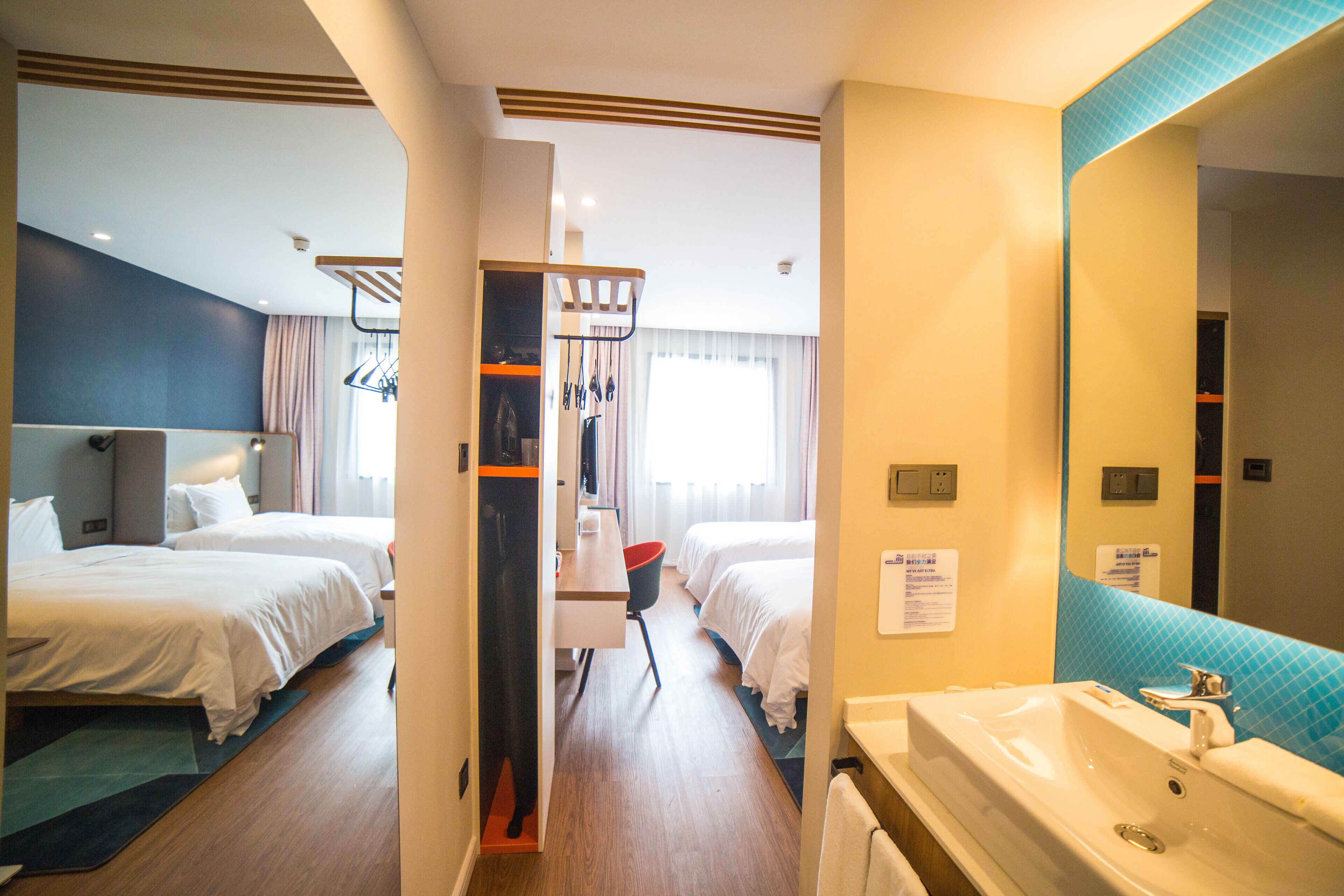 A hotel room with two beds, a desk, a chair, a sink, and a mirror.