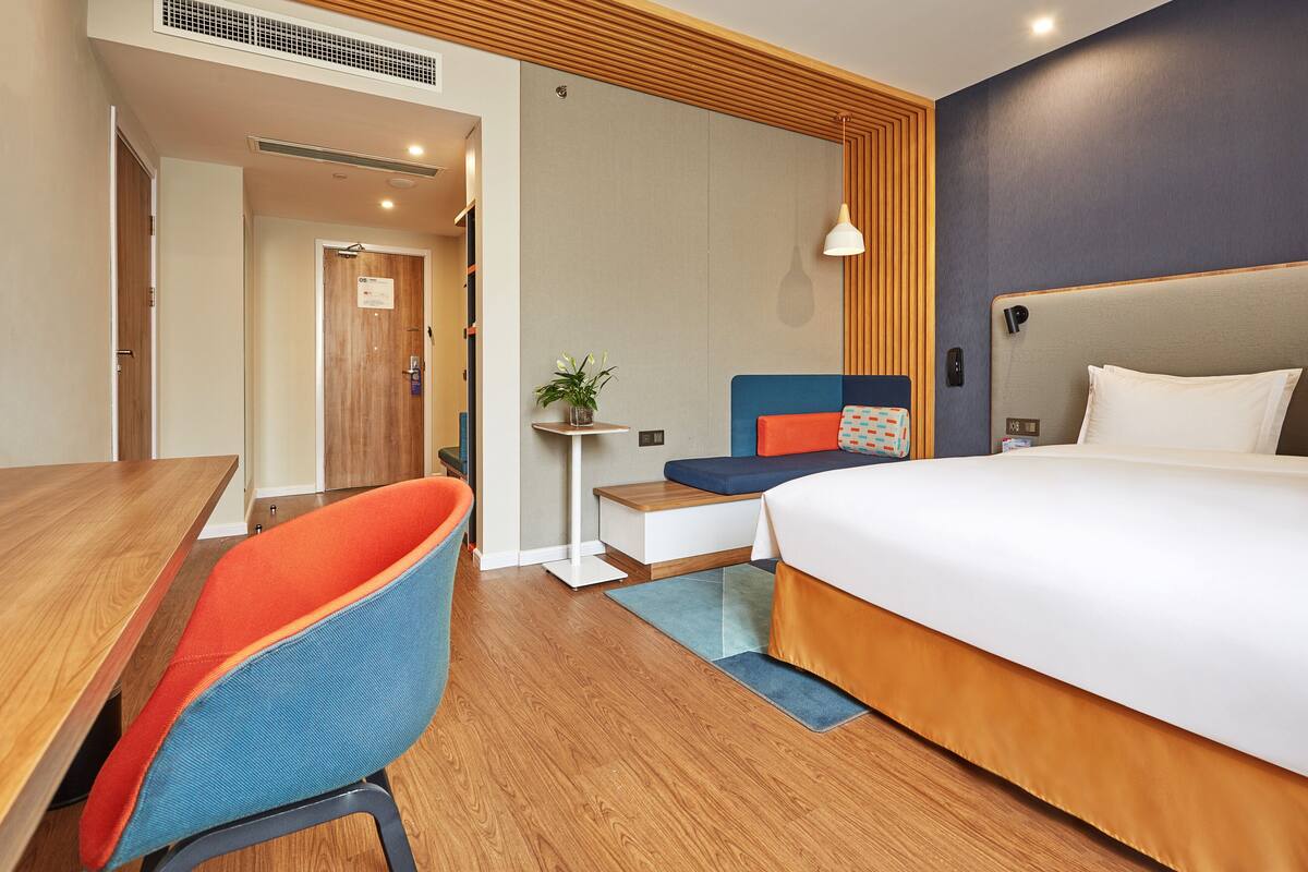 Ett hotellrum med ett skrivbord i trä, en blå och orange stol, en säng med vita och orange sängkläder och ett litet bord med en växt.
