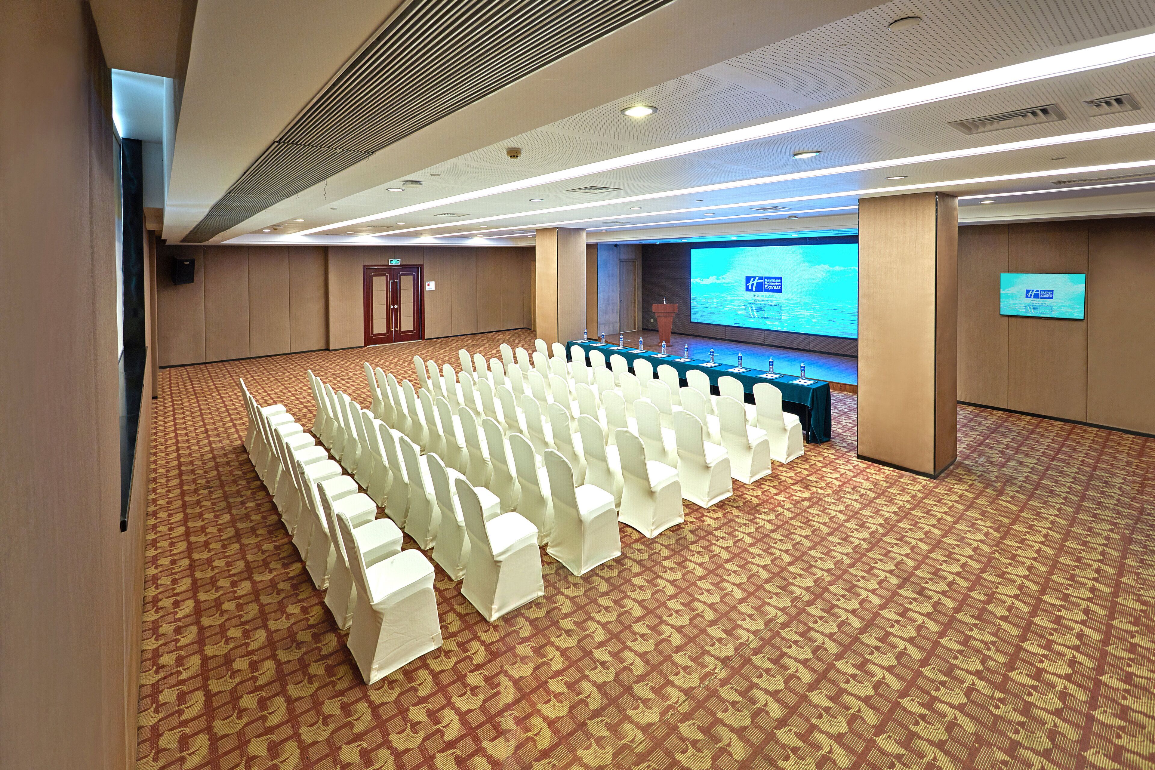 A conference room with rows of white chairs, a long table with microphones, and a large screen displaying a presentation.