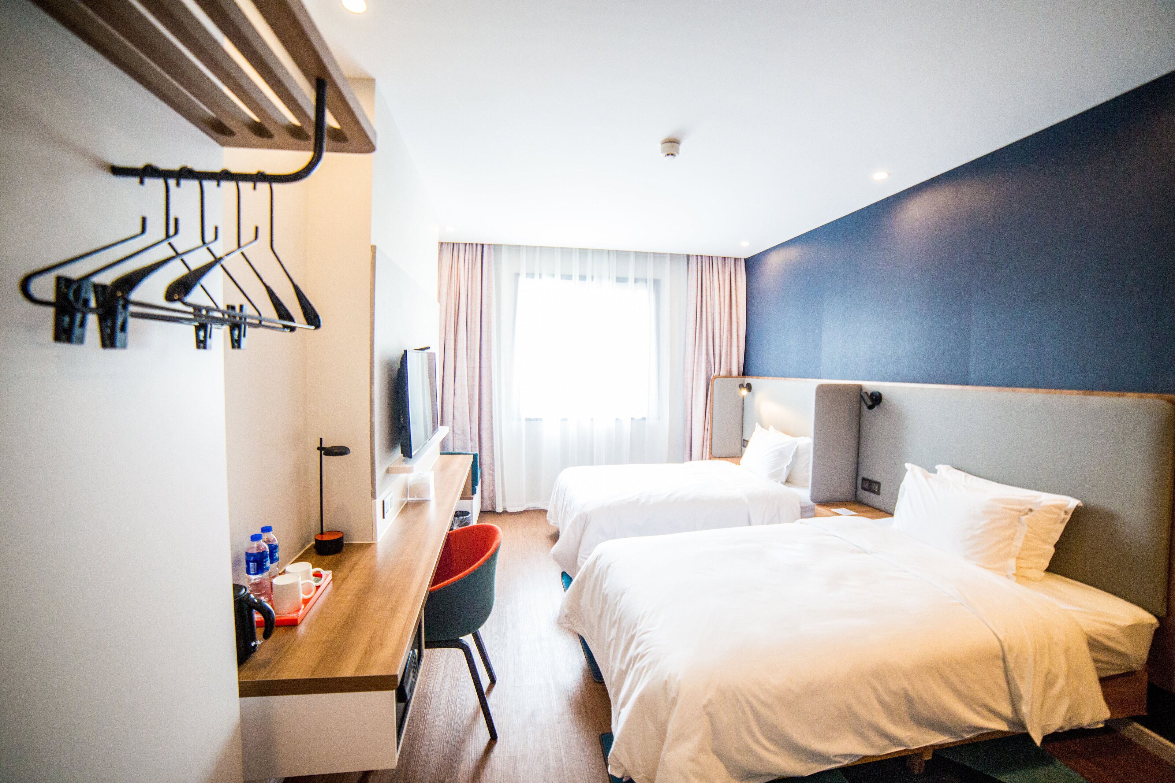 A hotel room with two beds, a desk with a chair, a TV, and a coat rack.
