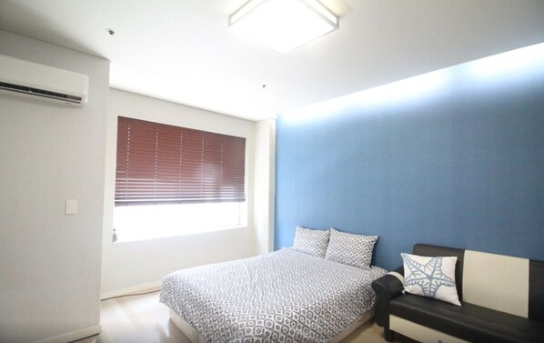 A bedroom with a bed, a window with blinds, a blue accent wall, and a small sofa. - GANGNAM SK Metro A (Seoul)