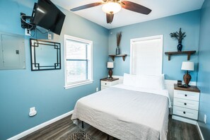 A bedroom with a bed, nightstands, a TV mounted on the wall, a ceiling fan, and a window with blinds.