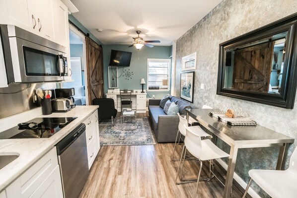 A compact kitchen with a microwave, toaster, and dishwasher. There is a dining area with a table and chairs, and a sofa. A ceiling fan is visible.