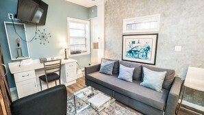 A modern living room with a grey sofa, a glass coffee table, a desk with a chair, and a television mounted on the wall. - Inn The Heights - Apt. 9 (Tampa)