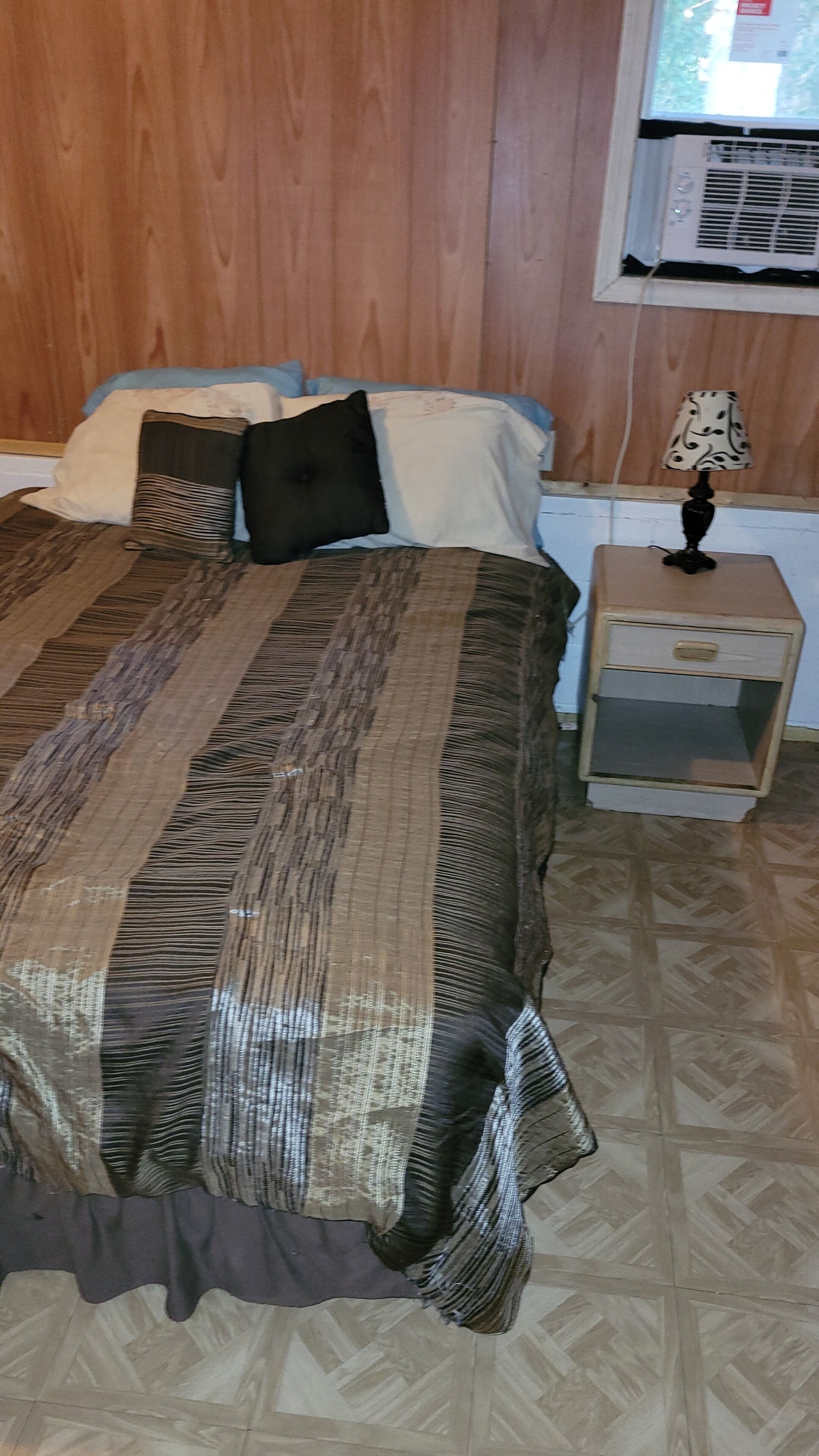 A bed with a patterned bedspread, a bedside table with a lamp, and a window with an air conditioner.