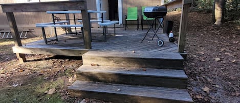 A wooden deck with picnic tables and a barbecue grill, leading to a building with a door and windows.