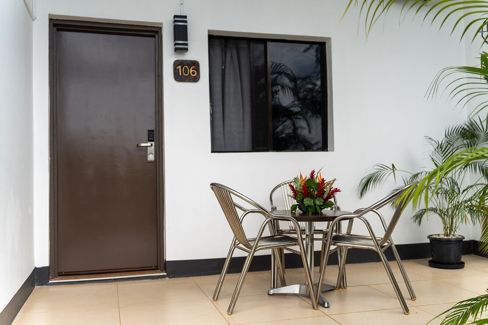 Una habitación con puerta cerrada, ventana con vista a palmeras, una mesa con sillas y una planta en maceta.