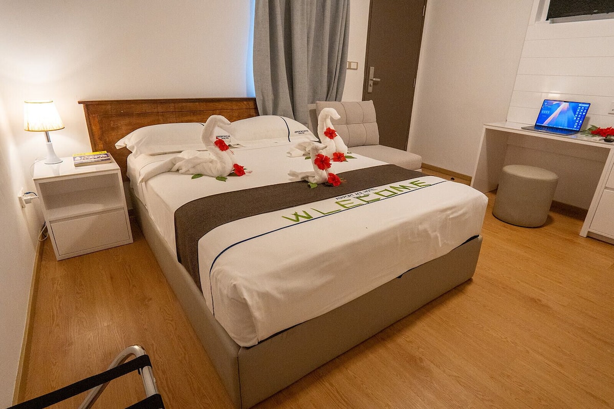 A neatly made bed with a welcome mat, a bedside lamp, a laptop on a desk, and a small trash bin.