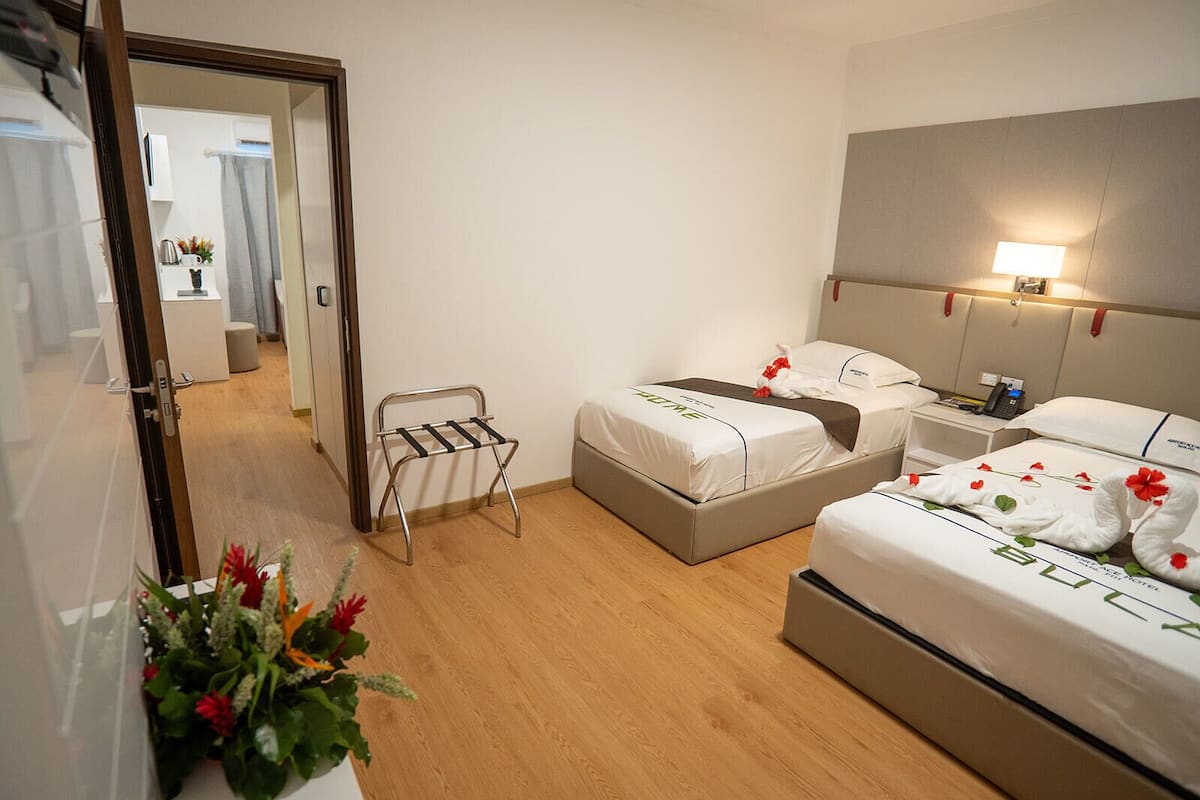 A hotel room with two beds, a bedside table, a lamp, and a small stool.