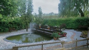 Um jardim tranquilo com um pequeno lago, uma ponte de madeira e diversos vasos de plantas.