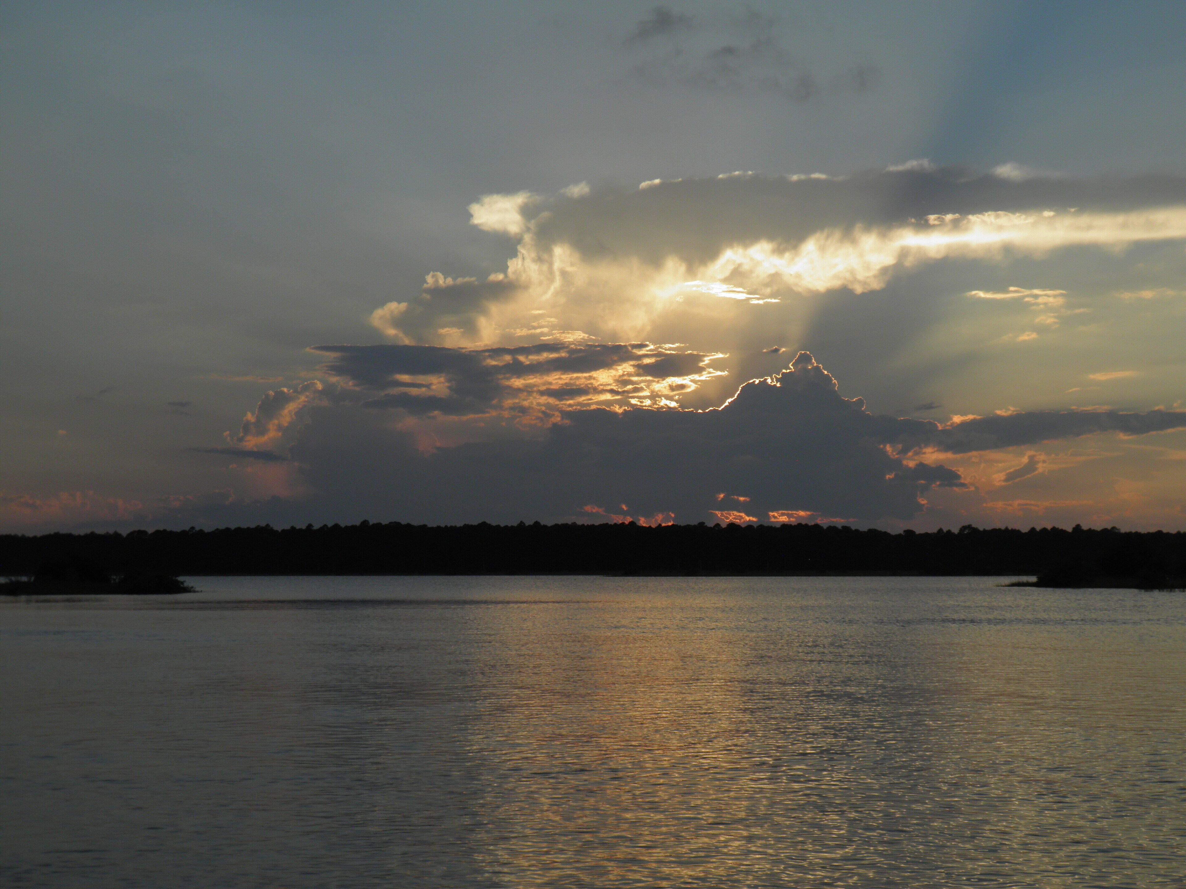 A sunset over a calm body of water with a silhouette of a forested landmass in the distance.