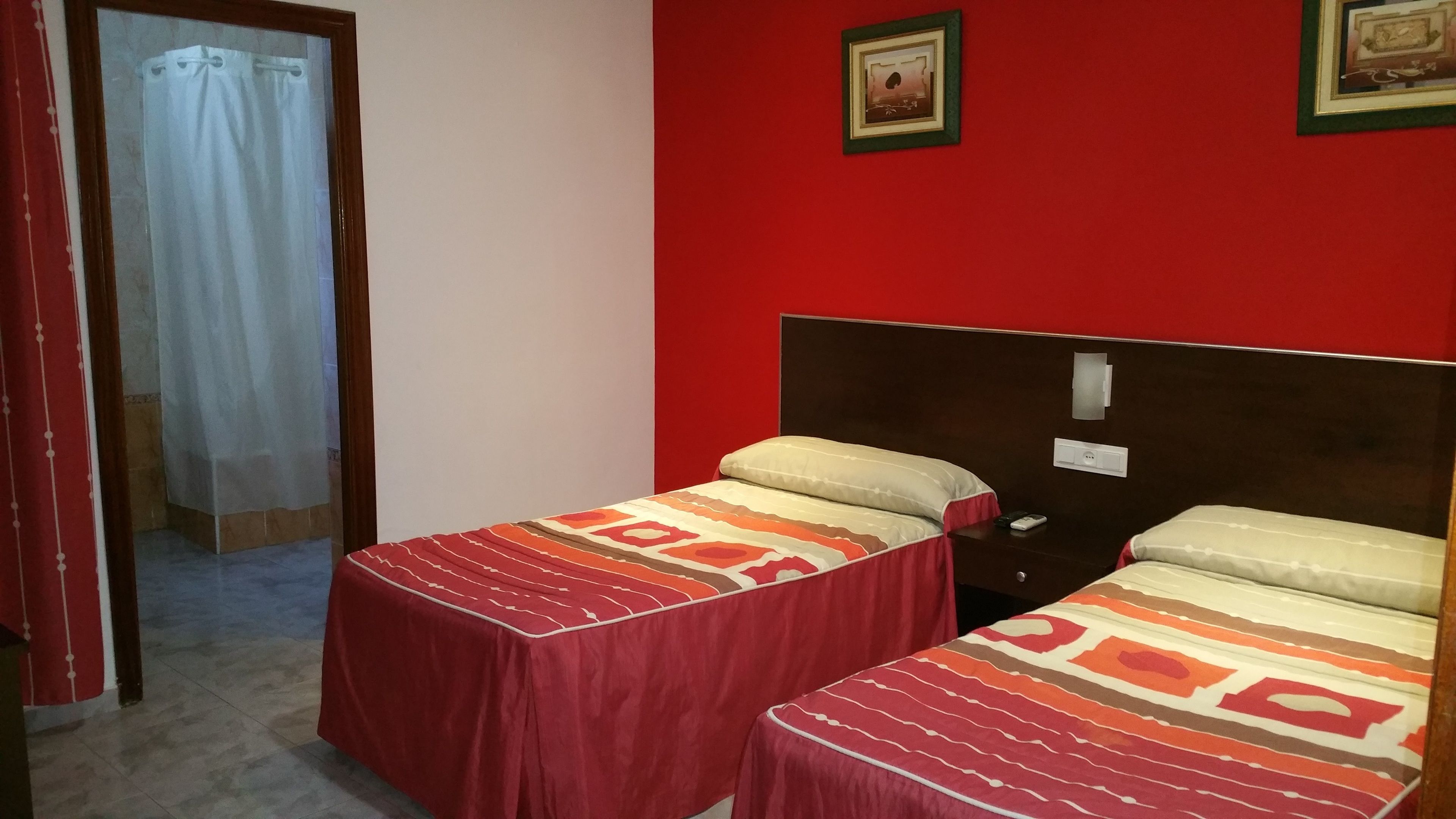 Habitación con dos camas, cada una con colcha y almohadas estampadas. Tiene un cabecero de madera, una mesita con lámpara y un interruptor de luz en la pared.