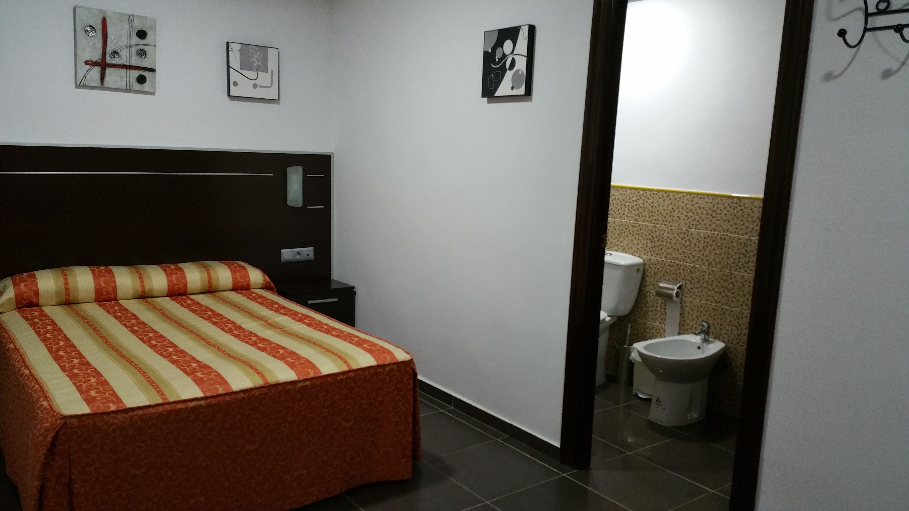 A hotel room with a bed, a nightstand, a wall-mounted light, and a toilet visible through an open door.