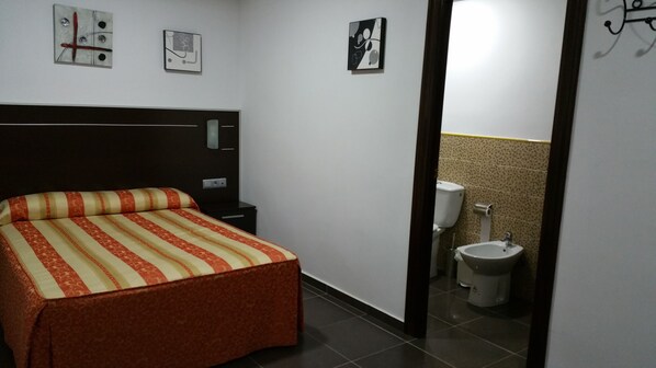Habitación de hotel con cama, mesita de noche, luz empotrada en la pared y un inodoro visible a través de una puerta entreabierta.