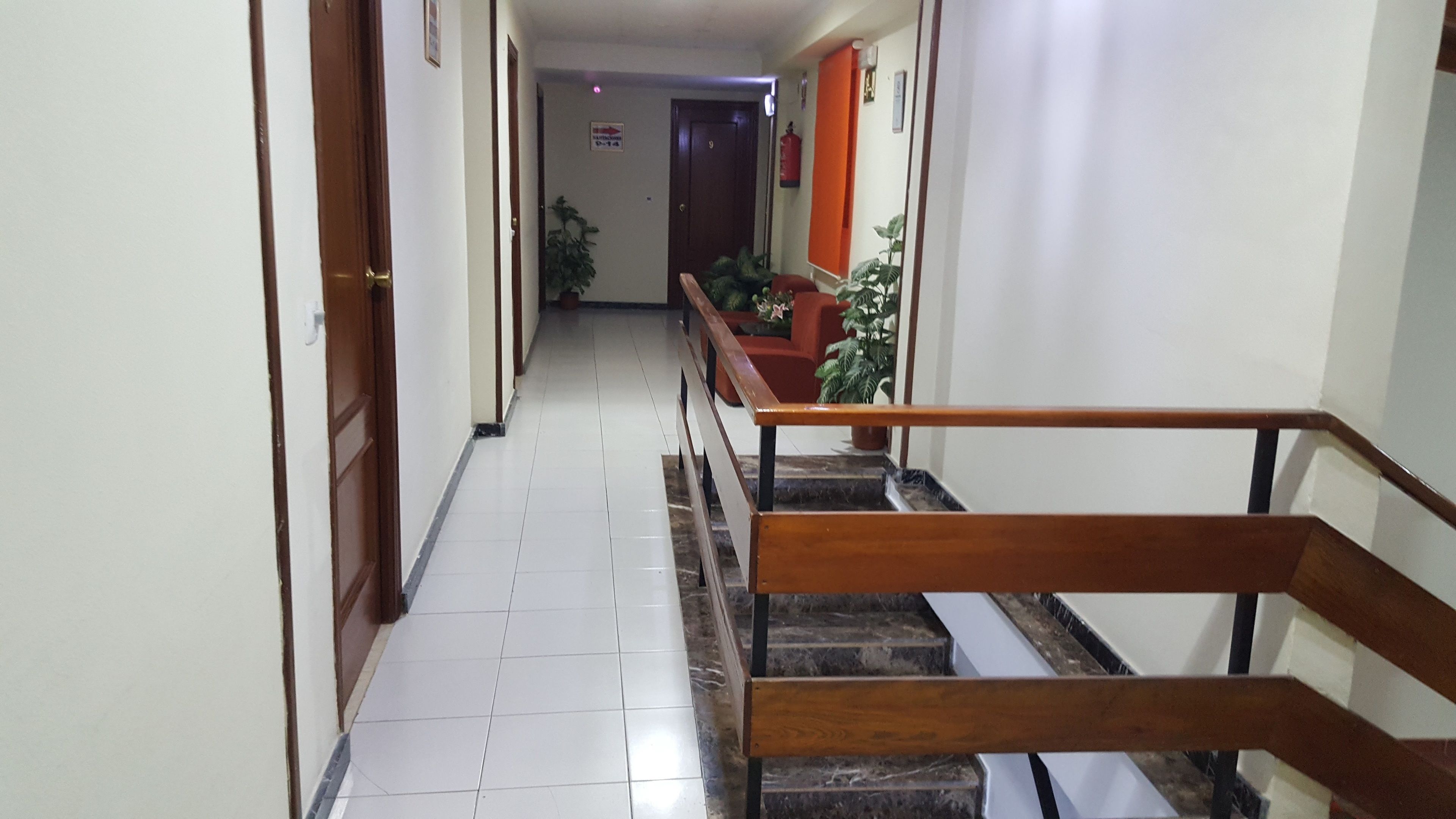 A hotel hallway with a wooden staircase, tiled floor, and potted plants.