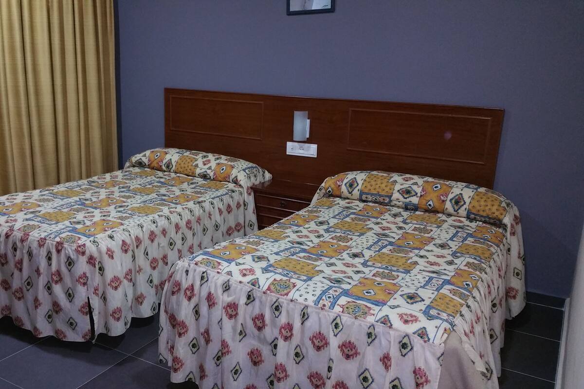 A room with two beds, each with a patterned bedspread, a wooden headboard, and a curtain on one side.