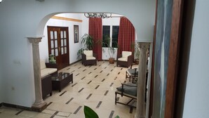 A spacious living area with a tiled floor, a sofa, two armchairs, a coffee table, and a dining table with chairs. There are potted plants and a large window with curtains. - Hostal Restaurante el Lirio (Bollullos Par del Condado)