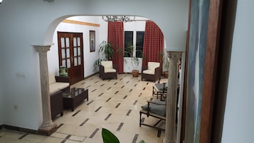 Amplia sala de estar con suelo de baldosas, un sofá, dos sillones, una mesa de centro y una mesa de comedor con sillas. Hay plantas en macetas y un ventanal con cortinas.