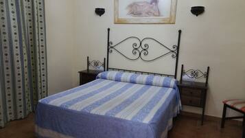 Un dormitorio con cama, mesita de noche, una silla, cortina y un cuadro en la pared.