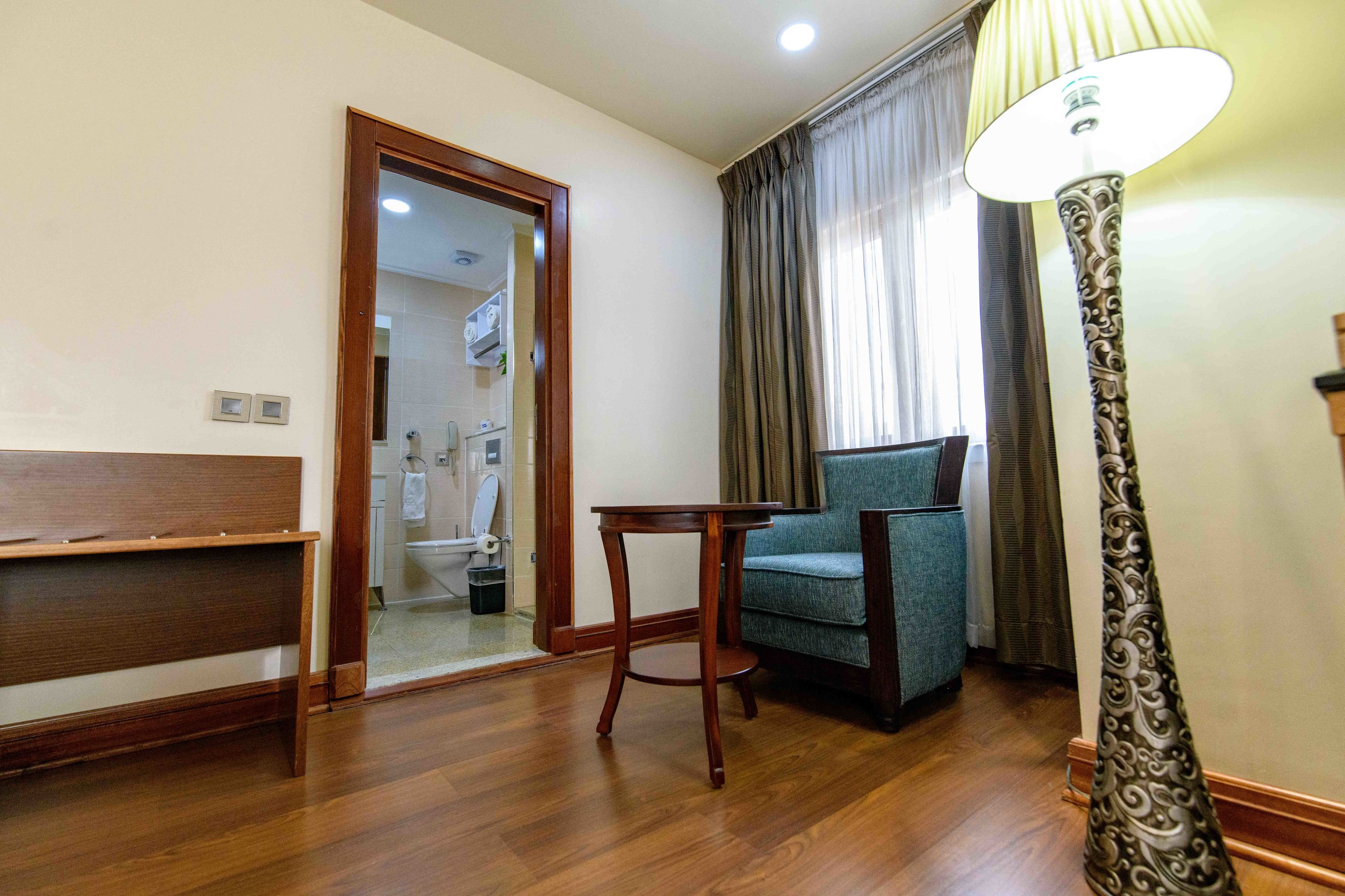 Una habitación de hotel con un banco de madera, una mesita pequeña, un sillón azul y una lámpara de pie.