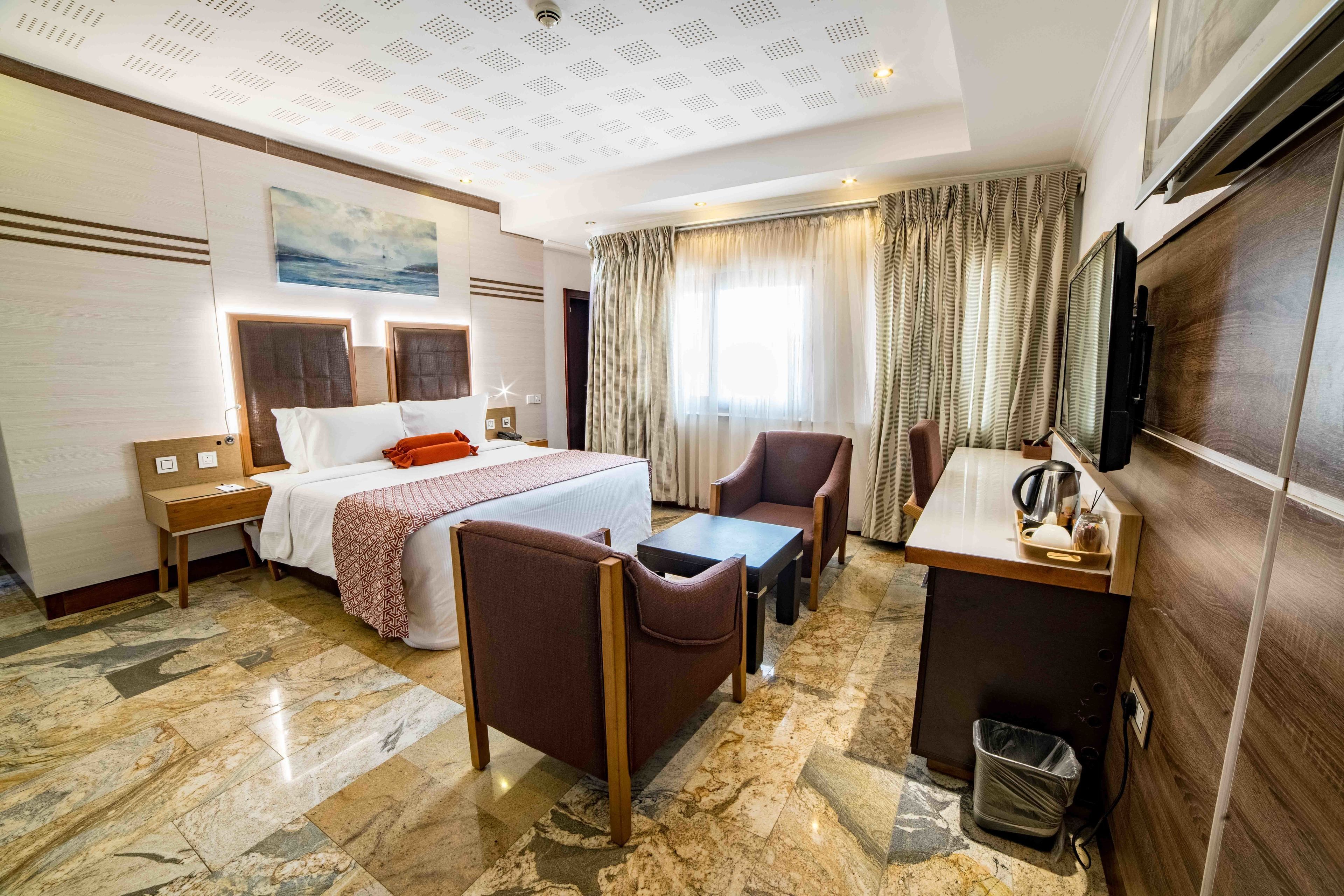 A hotel room with a bed, two chairs, a small table, a TV, and a desk with a kettle.