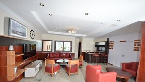 Uma sala ampla com um sofá vermelho, uma mesa de centro de madeira e uma televisão. Há uma cozinha com uma geladeira e uma pia.