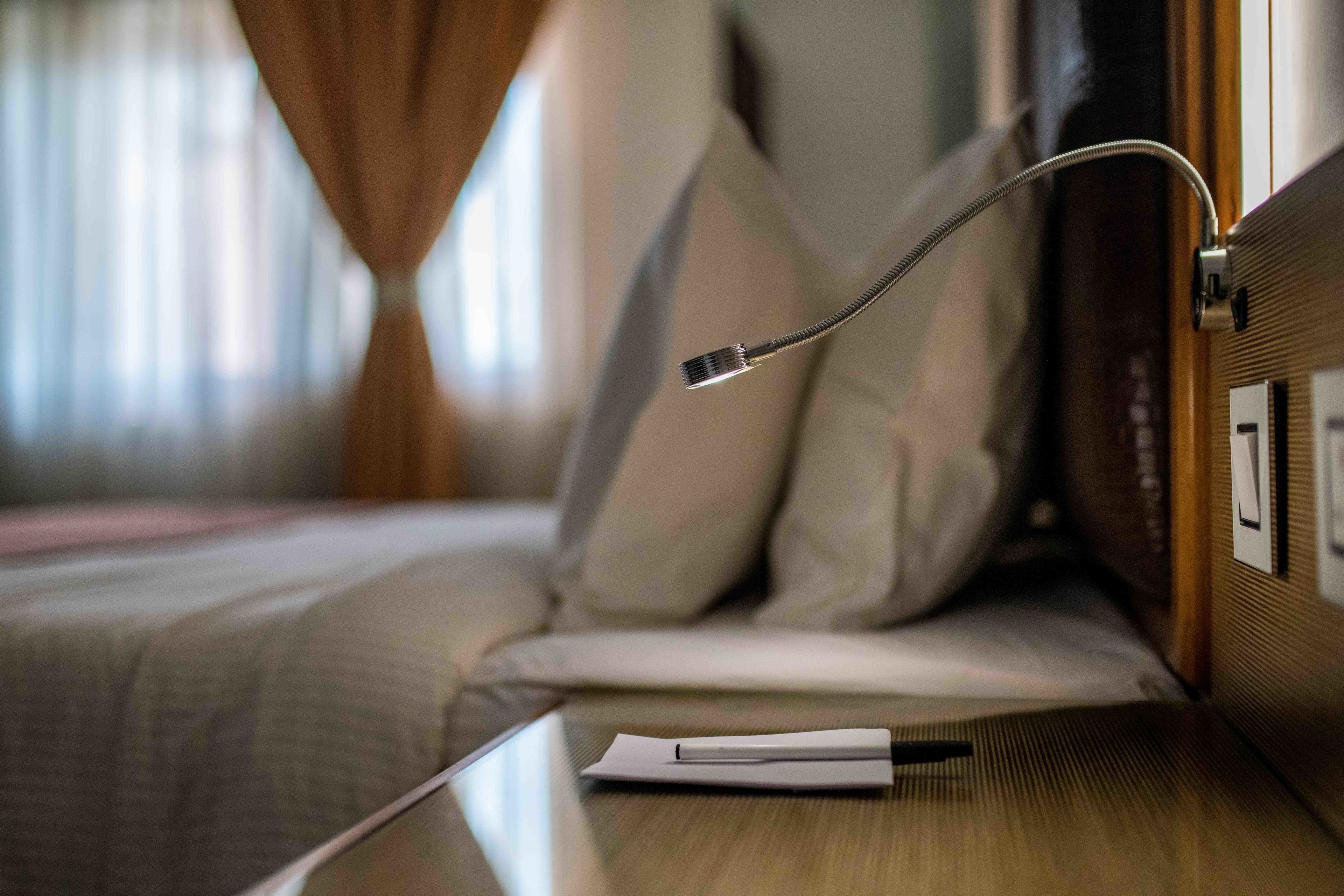 A neatly made bed with a white comforter and pillows, a bedside table with a lamp, and a window with curtains.