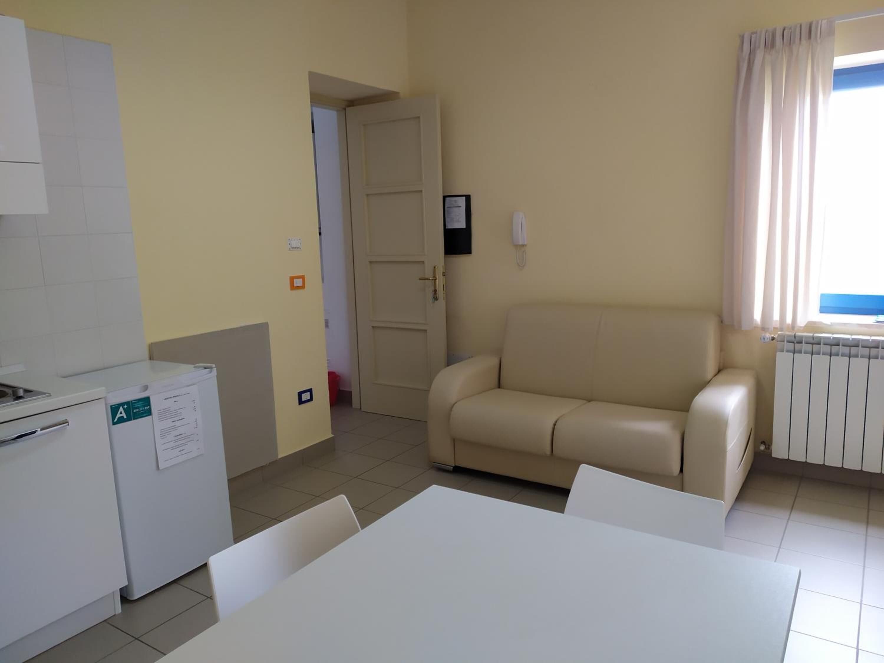 A compact room with a kitchenette, a beige sofa, a dining table, and a window with curtains.