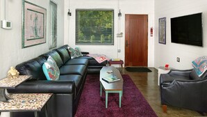 A living room with a black leather sofa, a coffee table, a ceiling fan, and a large window with blinds. - Folly Vacation 204 Getaway, Relax & Unwind (Folly Beach)
