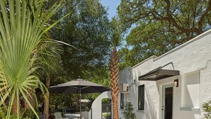 An outdoor patio with a table and chairs, surrounded by palm trees and a white building with a door and windows. - Folly Vacation 204 Getaway, Relax & Unwind (Folly Beach)