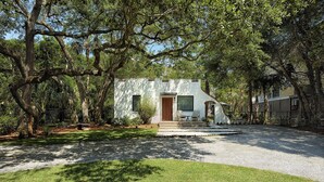 A small, white building with a wooden door and windows, surrounded by lush greenery and trees. - Folly Vacation 204 Getaway, Relax & Unwind (Folly Beach)